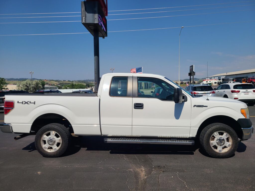 White Ford F-150 pickup with 4x4 badge parked in a lot, American flag mounted on the cab against a clear blue sky.