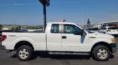 White Ford F-150 pickup with 4x4 badge parked in a lot, American flag mounted on the cab against a clear blue sky.