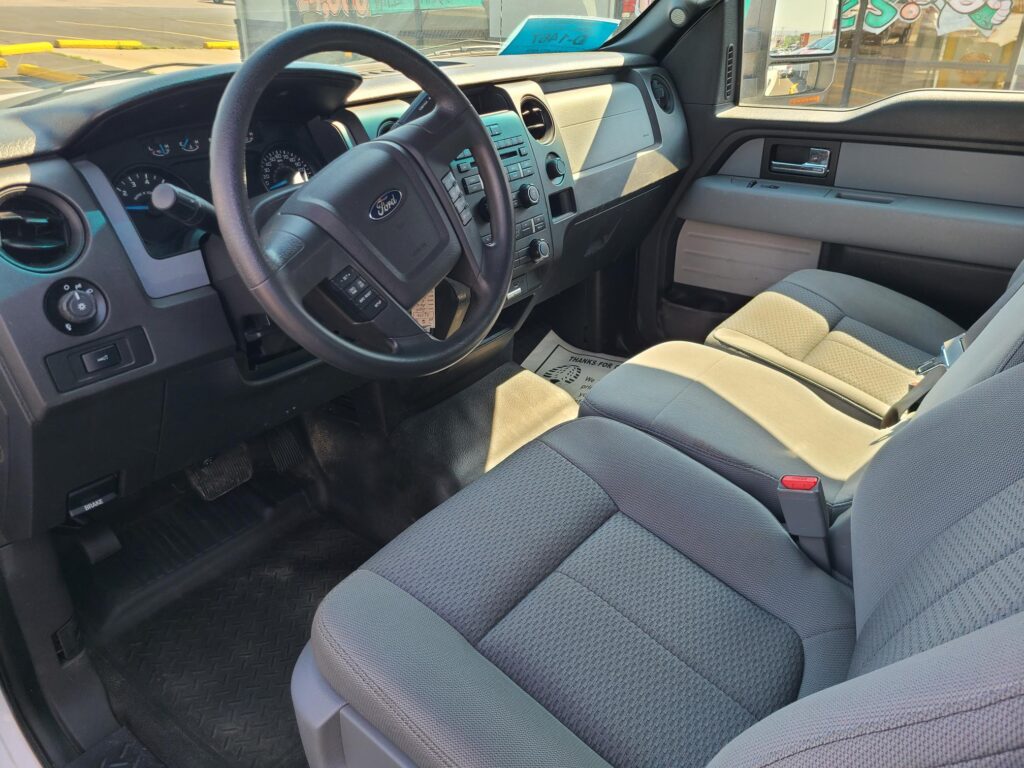 Interior view of a Ford truck cabin showing the steering wheel, instrument cluster, and center console with many buttons; gray fabric seats and sunlit dashboard.