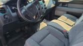 Interior view of a Ford truck cabin showing the steering wheel, instrument cluster, and center console with many buttons; gray fabric seats and sunlit dashboard.