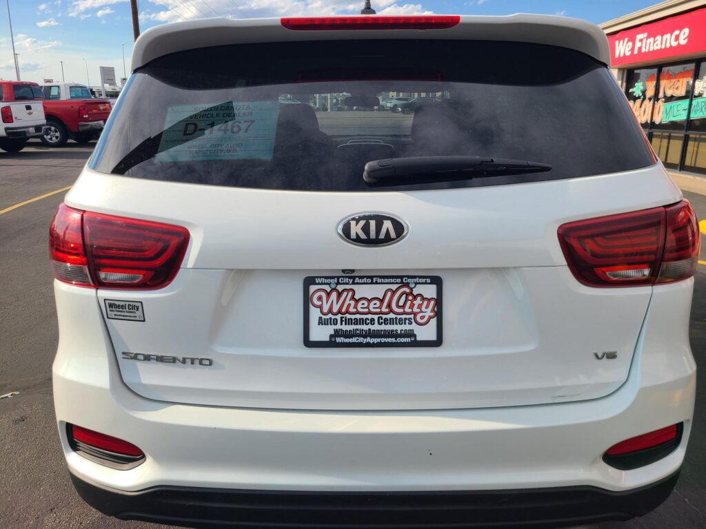 Rear view of a white Kia Sorento in a dealership lot; visible dealer plate frame and a window sticker showing D-1467, with a 'We Finance' sign in the background.