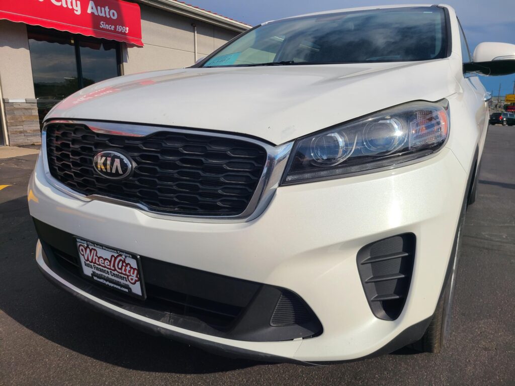 White Kia SUV parked at a Wheel City Auto dealership, showing the front grille and headlight with a windshield sticker visible.