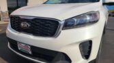 White Kia SUV parked at a Wheel City Auto dealership, showing the front grille and headlight with a windshield sticker visible.