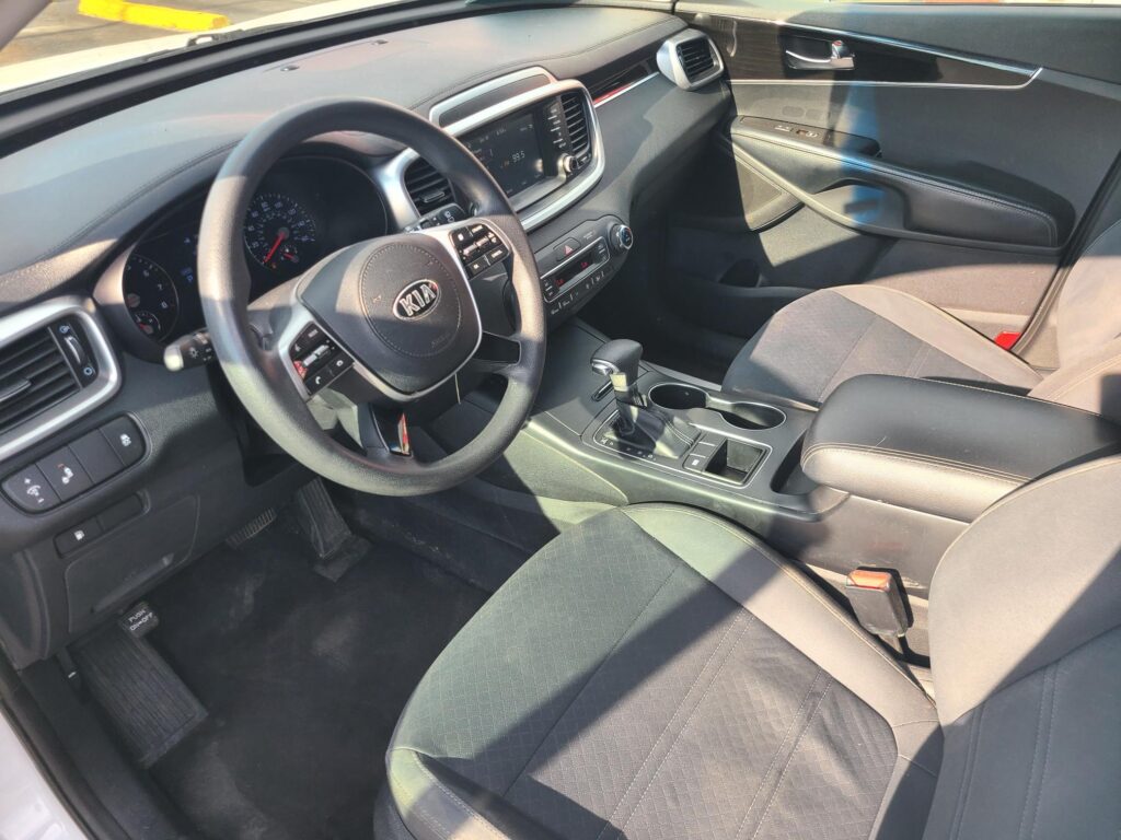 Interior view of a Kia sedan: steering wheel with Kia logo, center touchscreen, climate controls, and black fabric seats ahead of the driver.