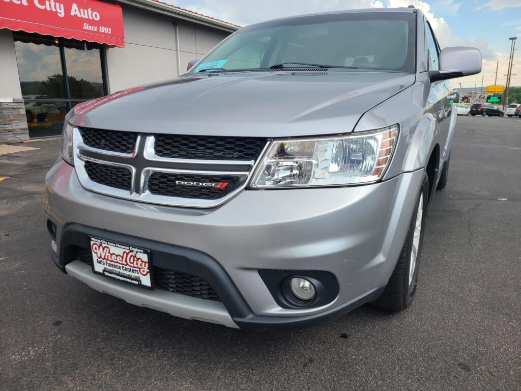 Silver Dodge minivan parked at a dealership, front view with Wheel City Auto license plate frame and red awning in the background