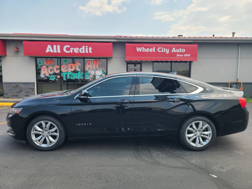 Black sedan parked in front of a car dealership with red signs reading All Credit and Wheel City Auto