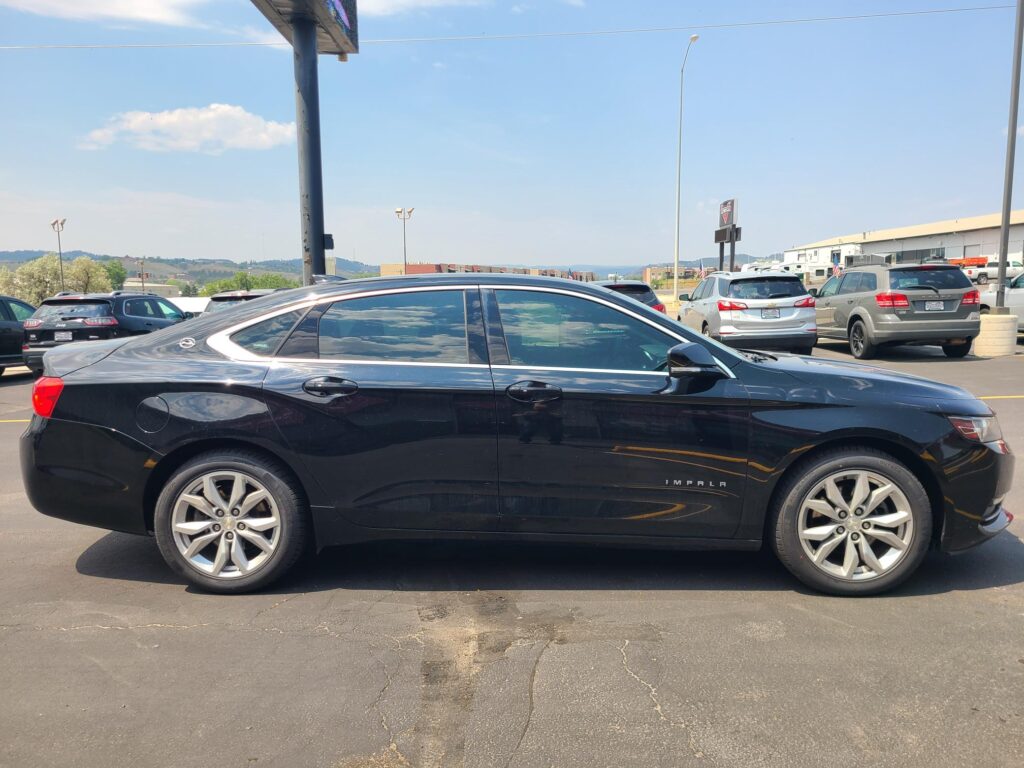 Close-up of a black Chevrolet Impala in a parking lot, with other cars and a light industrial building in the background under a blue sky.