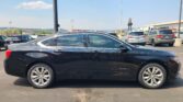 Close-up of a black Chevrolet Impala in a parking lot, with other cars and a light industrial building in the background under a blue sky.