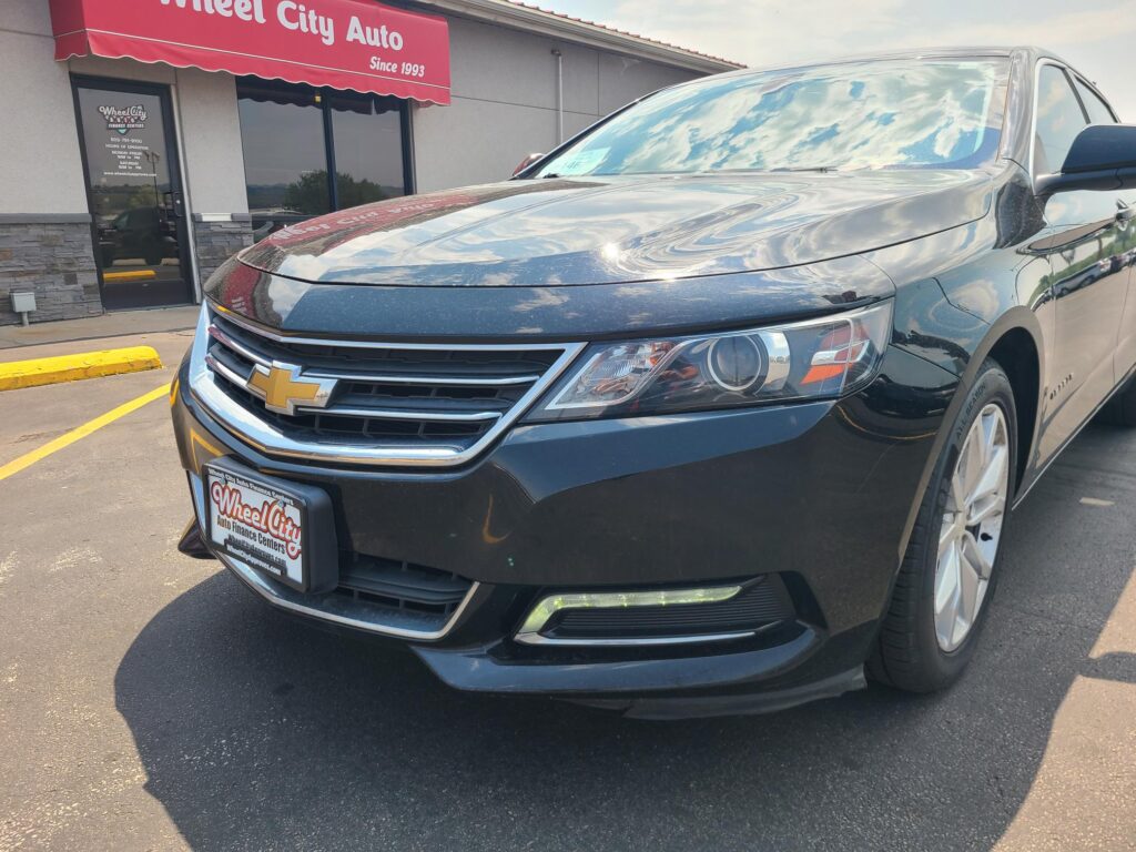 Front of a dark gray Chevrolet SUV parked in a lot with a red Wheel City Auto storefront sign in the background