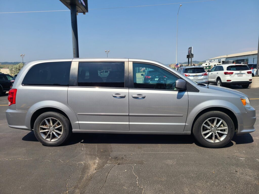 Silver minivan in the foreground of a car dealership lot, with other vehicles and a building in the background on a clear, sunny day.