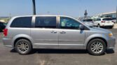 Silver minivan in the foreground of a car dealership lot, with other vehicles and a building in the background on a clear, sunny day.