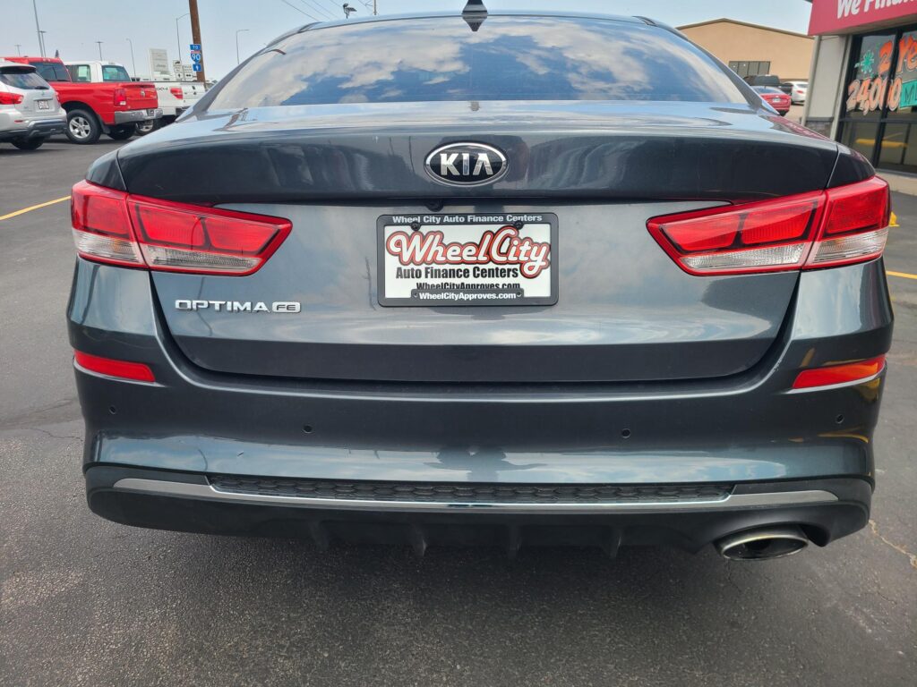 Rear view of a gray Kia Optima with a Wheel City Auto Finance Centers license plate frame in a dealership parking lot