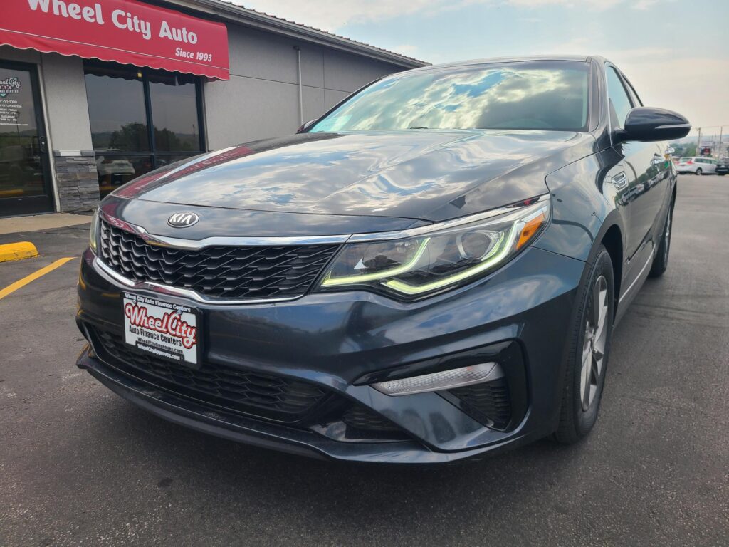 Front-right view of a dark gray Kia SUV parked outside a dealership with a red Wheel City Auto sign and license plate frame visible