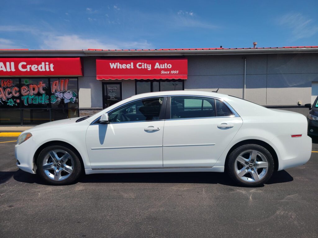 White sedan parked in front of a gray auto shop with a red Wheel City Auto awning and clear blue sky above.