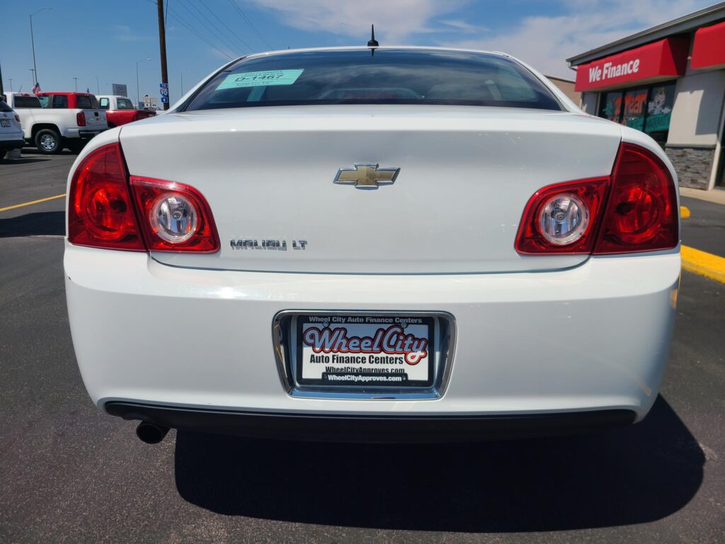 Rear view of a white Chevrolet Malibu LT in a dealership lot, showing the red taillights and Chevy bowtie emblem with Wheel City license plate frame in the foreground.
