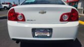 Rear view of a white Chevrolet Malibu LT in a dealership lot, showing the red taillights and Chevy bowtie emblem with Wheel City license plate frame in the foreground.