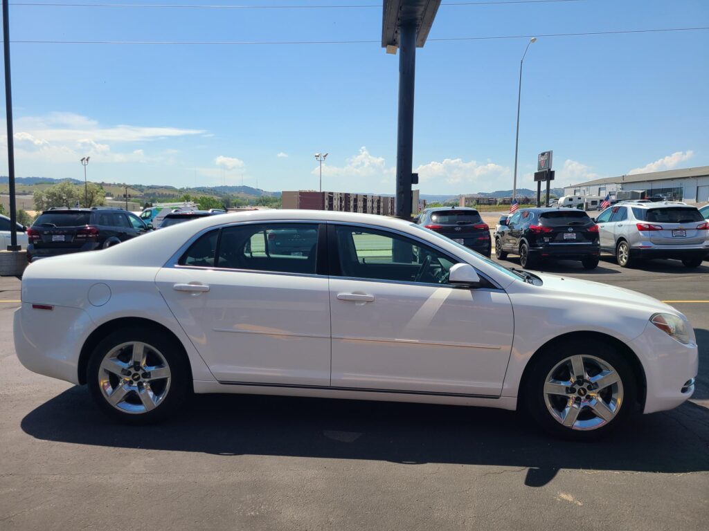 White sedan in the foreground of a crowded parking lot with other cars, a clear blue sky and distant hills.