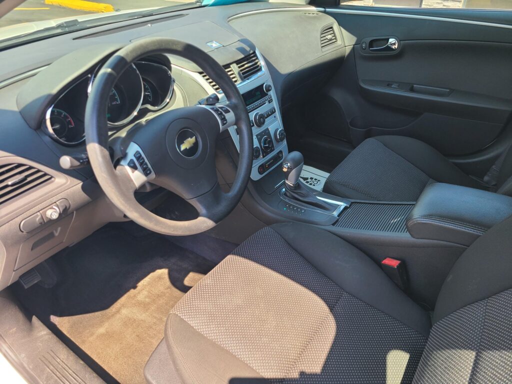Interior of a Chevrolet car showing the steering wheel with the Chevy emblem, center console, and front seats viewed from the driver’s side.]","Driver’s cockpit with dashboard, air vents, and gear shifter in a dark fabric interior.