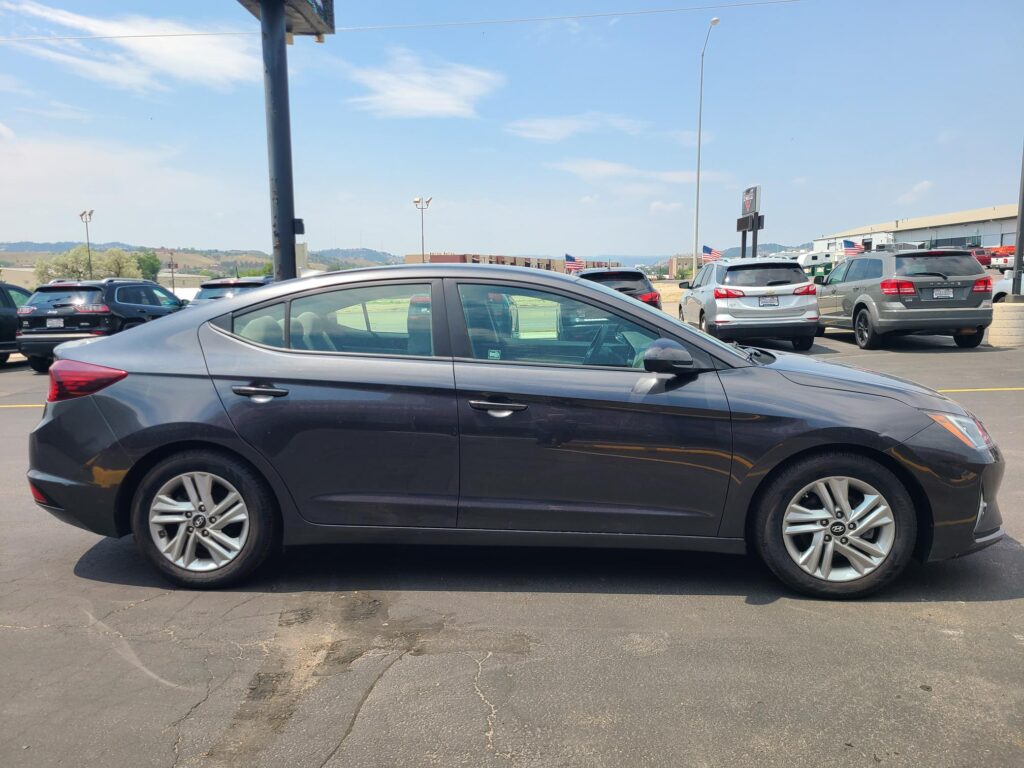 Gray Hyundai sedan in foreground in a crowded parking lot on a sunny day with other cars and American flags in the background.