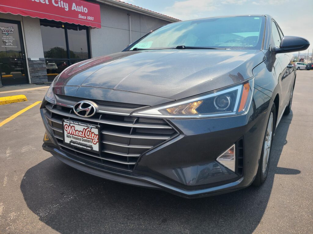 Front-left view of a gray Hyundai SUV parked in a dealership lot in front of Wheel City Auto with a red awning storefront.