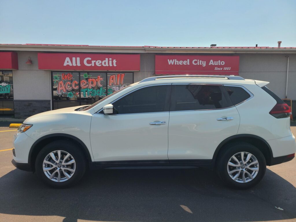 White SUV parked in front of a car dealership storefront with red 'All Credit' and 'Wheel City Auto' signs.