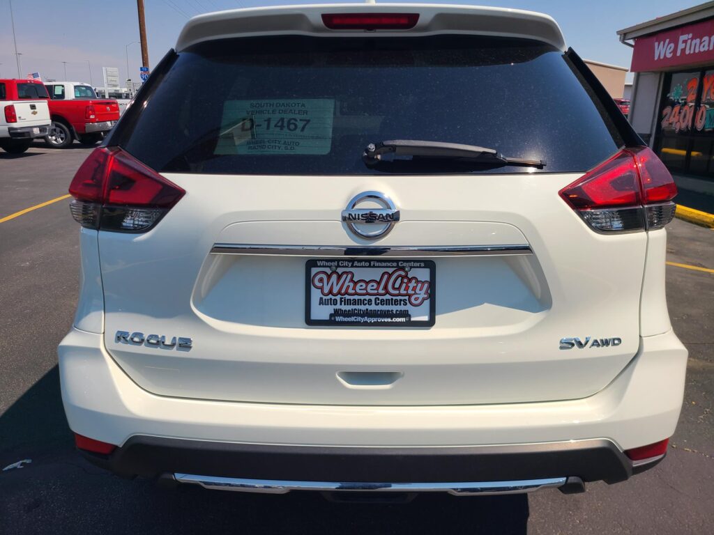 Rear view of a white Nissan Rogue SUV with Wheel City Auto Finance Centers dealer plate frame on the license plate area and a dusty rear window sticker.
