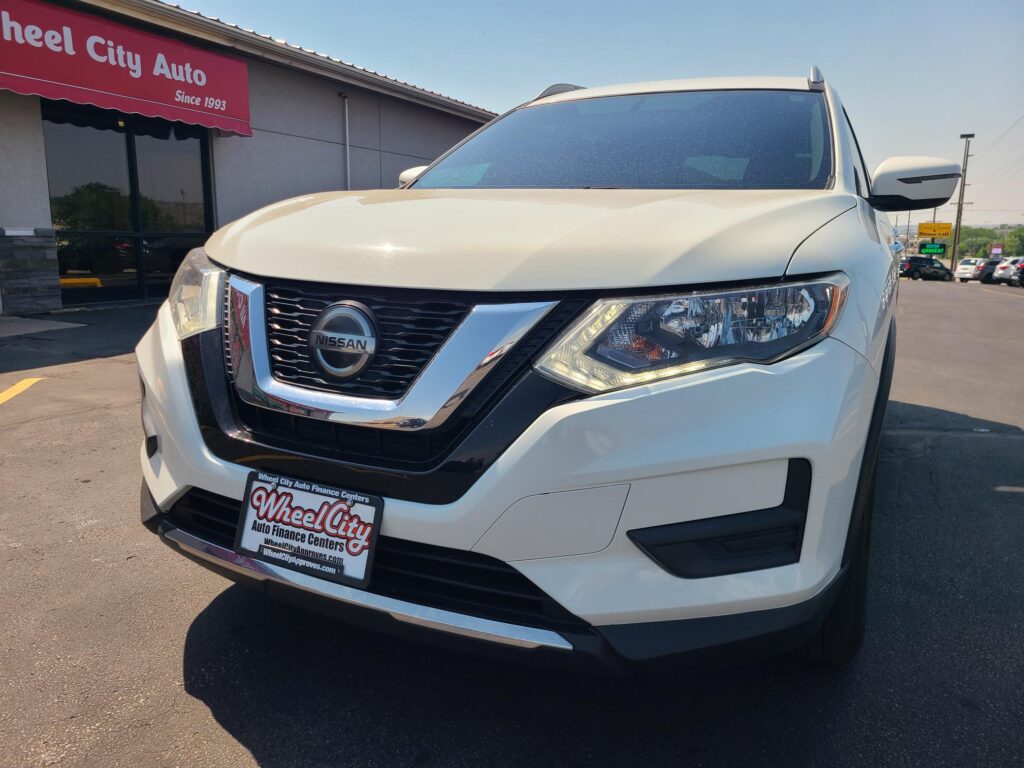 White Nissan SUV close-up at a dealership, Wheel City Auto Finance Centers sign visible in the background, front grille and logo in clear view.