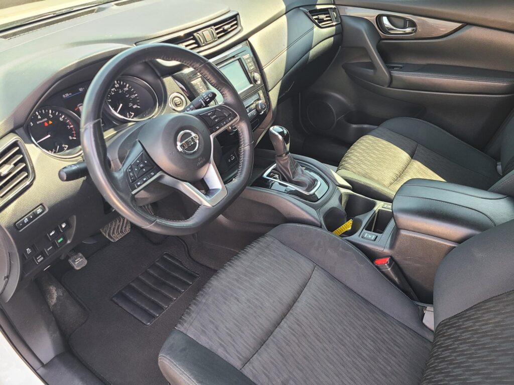Interior of a modern car showing the driver's seat, steering wheel, center console, and front passenger seat with sunlit cabin details.