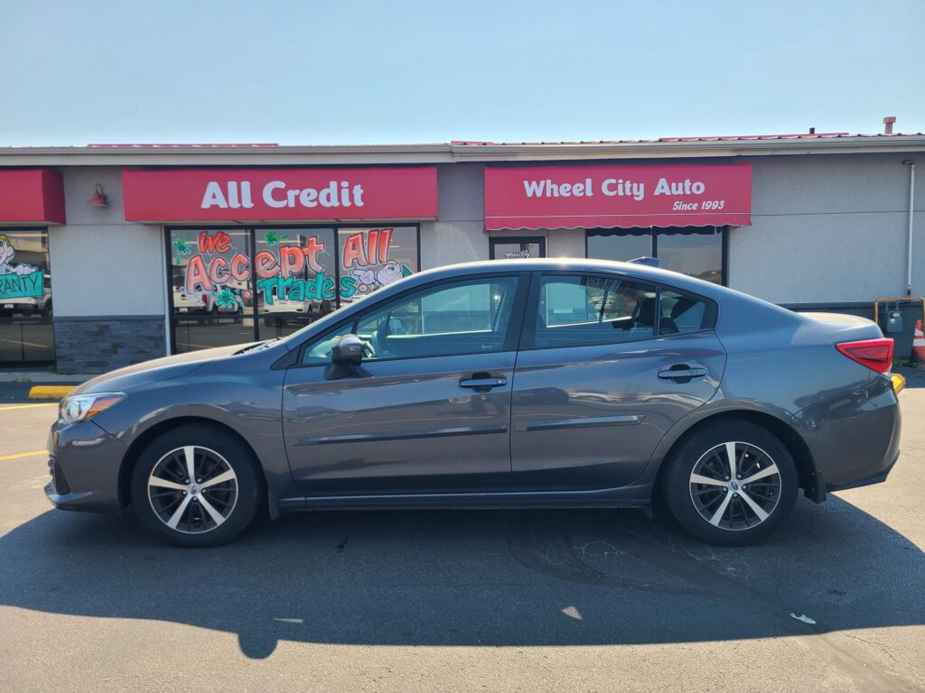 Gray sedan parked in a lot in front of a storefront with red signs reading All Credit and Wheel City Auto Auto Since 1993 .