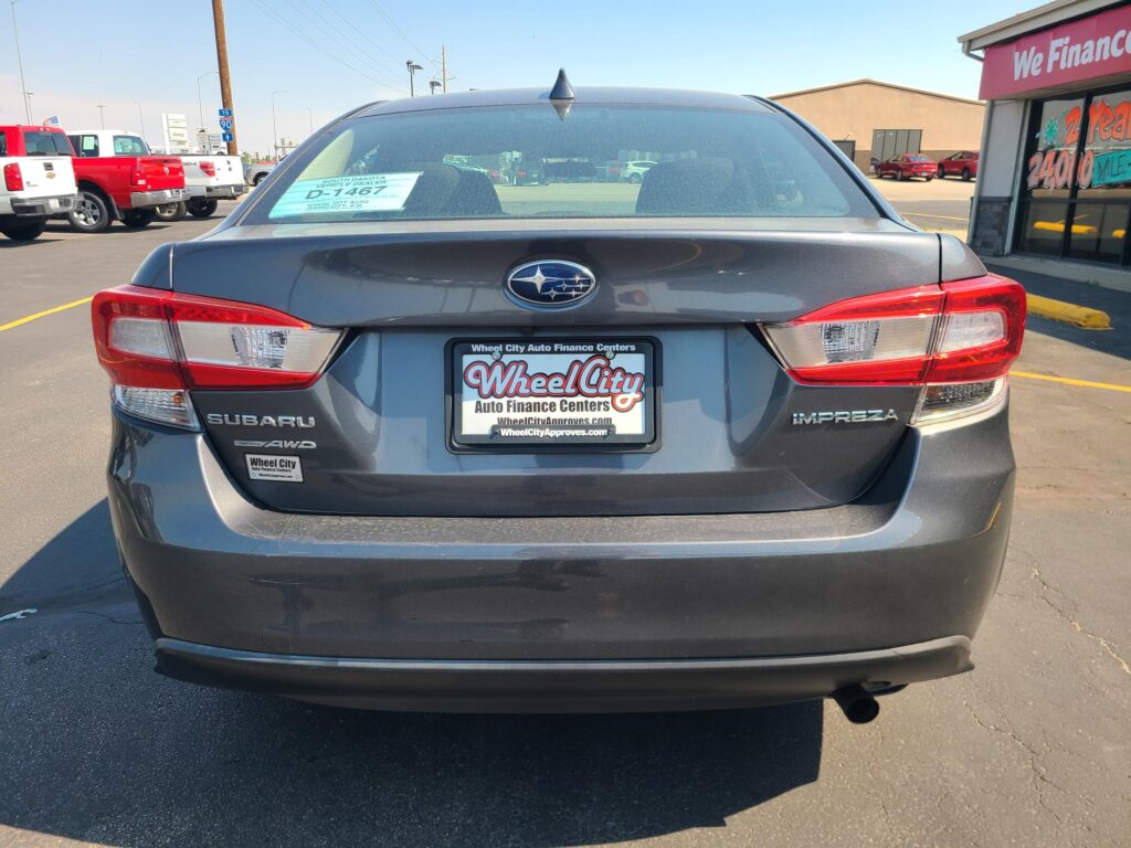 Rear view of a gray Subaru Impreza in a car lot, dealership plate reading WheelCity Auto Finance Centers, storefront in background.