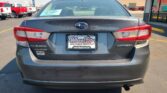 Rear view of a gray Subaru Impreza in a car lot, dealership plate reading WheelCity Auto Finance Centers, storefront in background.