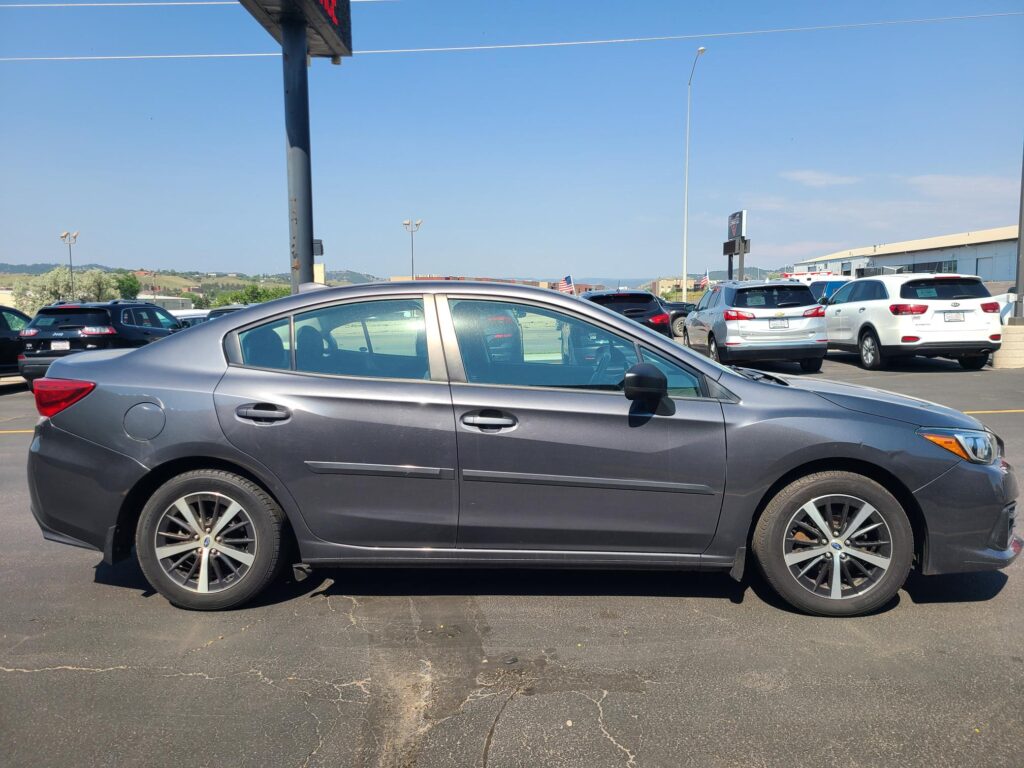 Side view of a dark gray sedan with alloy wheels in a crowded parking lot on a sunny day.