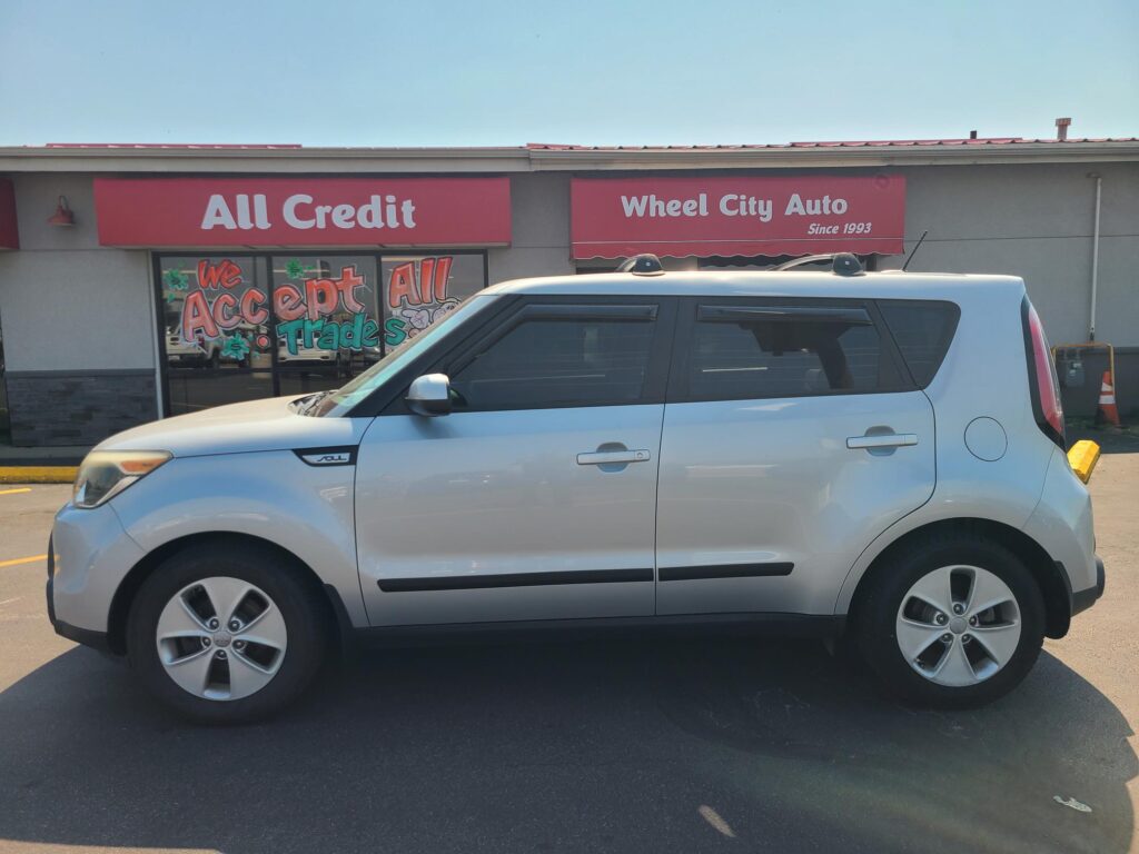 White SUV parked in front of a storefront with red All Credit and Wheel City Auto signs on the building. The window displays 'We Accept All Trades' promotional art.