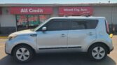 White SUV parked in front of a storefront with red All Credit and Wheel City Auto signs on the building. The window displays 'We Accept All Trades' promotional art.