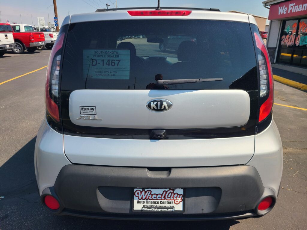 Rear view of a silver Kia Soul in a car dealership lot, with a Wheel City Auto Finance Centers license plate frame and dealer stickers on the rear window.