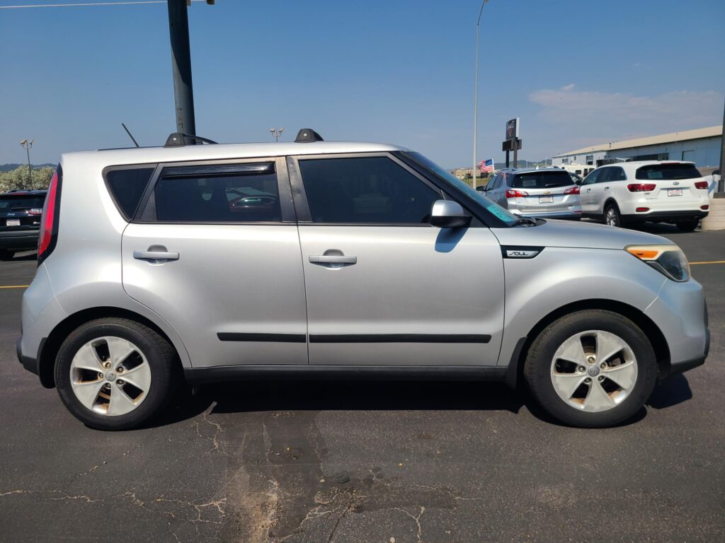 Side view of a silver Kia Soul SUV parked in a lot with other vehicles under a clear blue sky. A flag and dealership buildings are in the background.