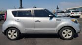 Side view of a silver Kia Soul SUV parked in a lot with other vehicles under a clear blue sky. A flag and dealership buildings are in the background.