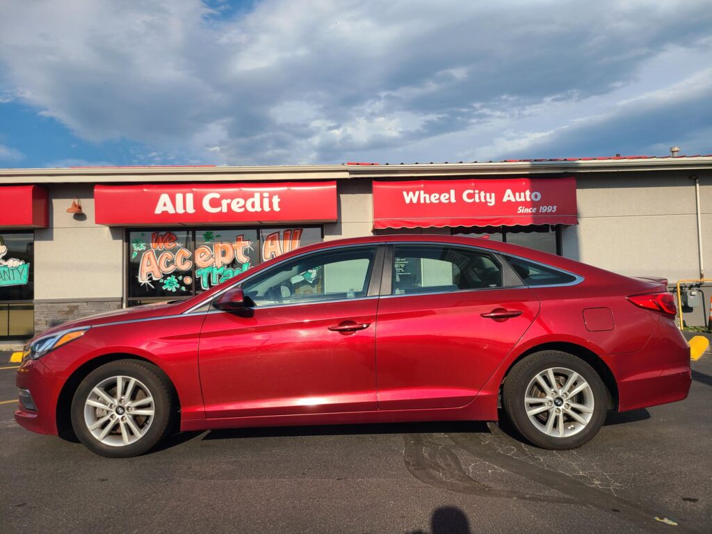 Red sedan parked in front of a storefront with bold red signs reading 'All Credit' and 'Wheel City Auto' under a cloudy sky.