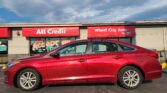 Red sedan parked in front of a storefront with bold red signs reading 'All Credit' and 'Wheel City Auto' under a cloudy sky.