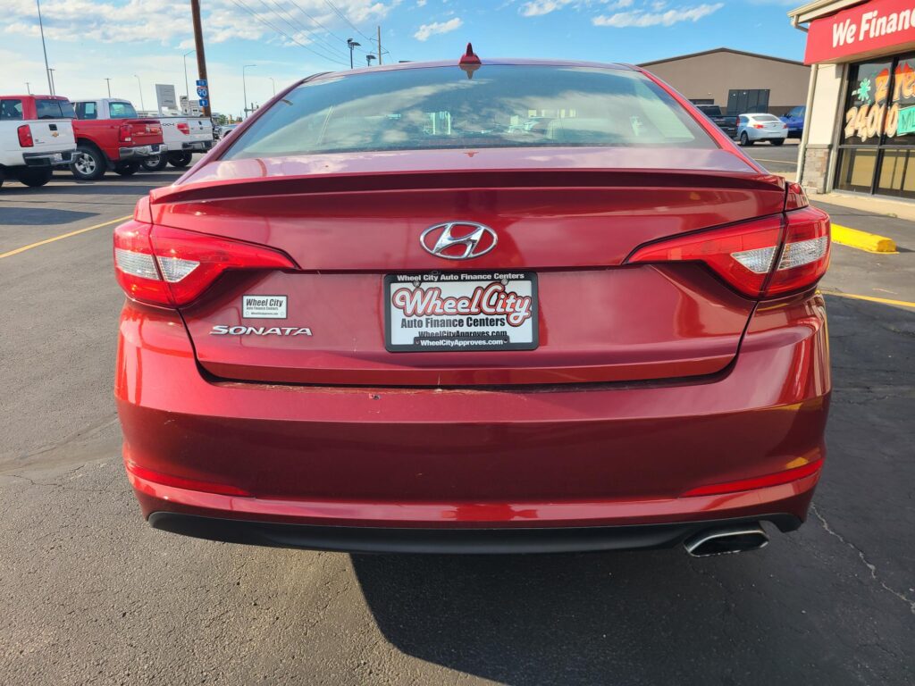 Rear view of a red Hyundai Sonata at a car dealership, license plate reads Wheel City Auto Finance Centers, with other trucks and a showroom on the right.