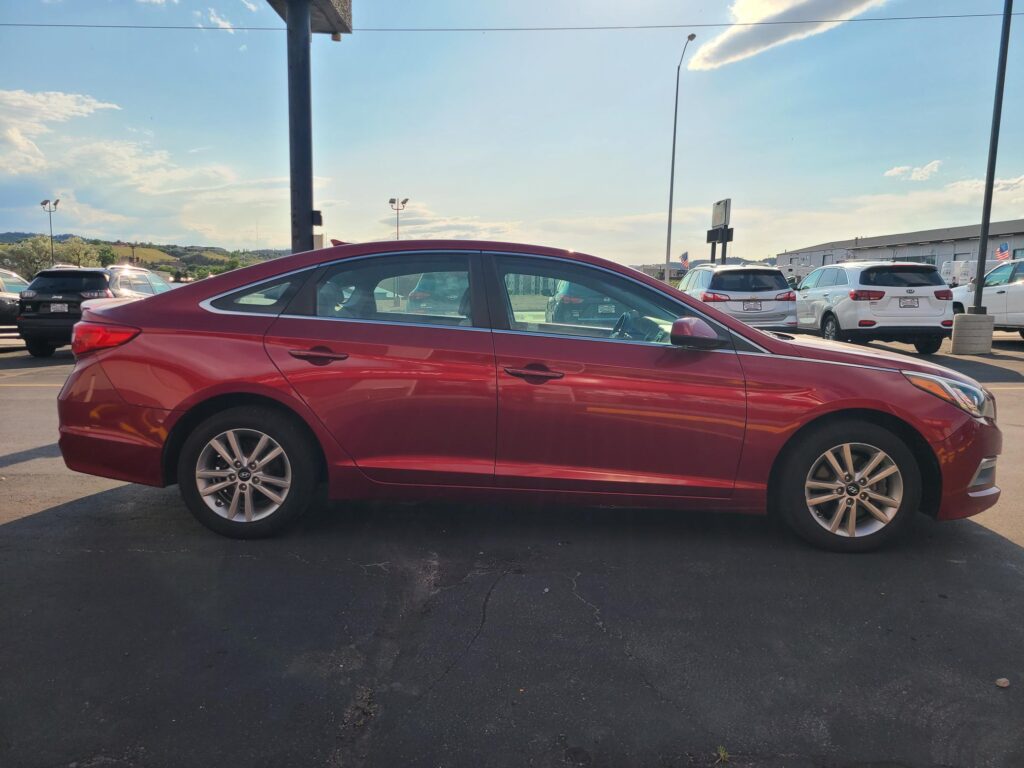 Red sedan parked in a lot, side view with other vehicles and blue sky in the background