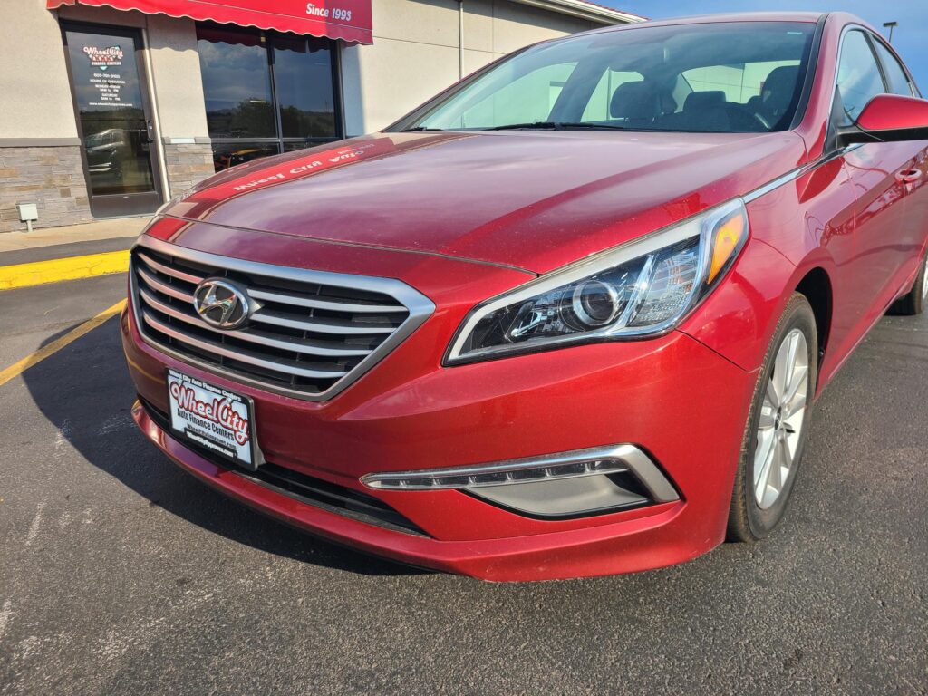 Red Hyundai minivan parked in front of Wheel City Auto Finance Centers storefront, tilted to show front grille and headlights.