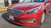 Red Hyundai minivan parked in front of Wheel City Auto Finance Centers storefront, tilted to show front grille and headlights.
