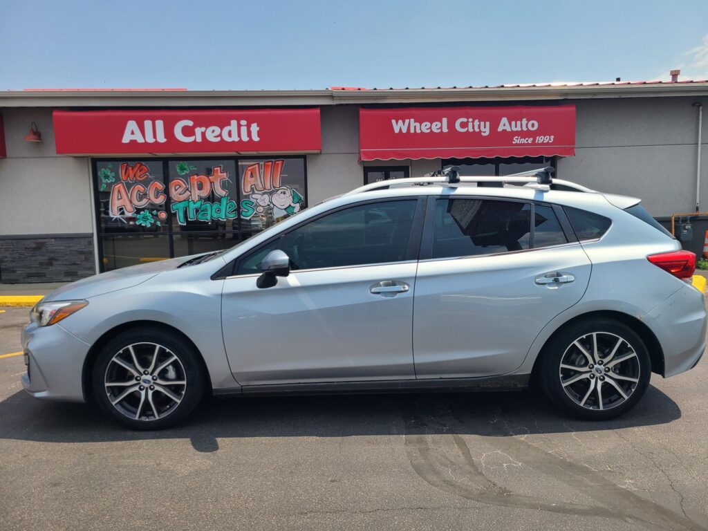 Front of a storefront with red signs reading 'All Credit' and 'Wheel City Auto Since 1993' and a silver SUV parked in front of the shop under a clear blue sky.