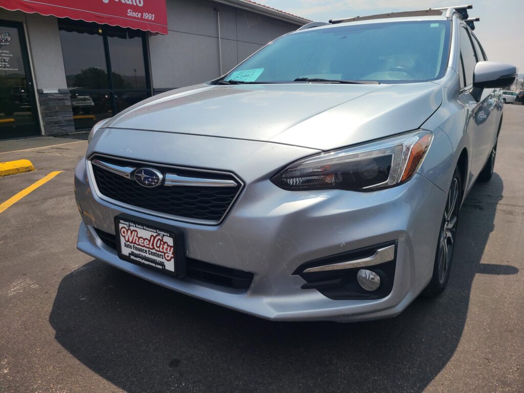 Front-left view of a silver Subaru SUV in a dealership lot, windshield sticker, and Wheel City license plate frame on the plate holder.