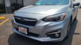 Front-left view of a silver Subaru SUV in a dealership lot, windshield sticker, and Wheel City license plate frame on the plate holder.