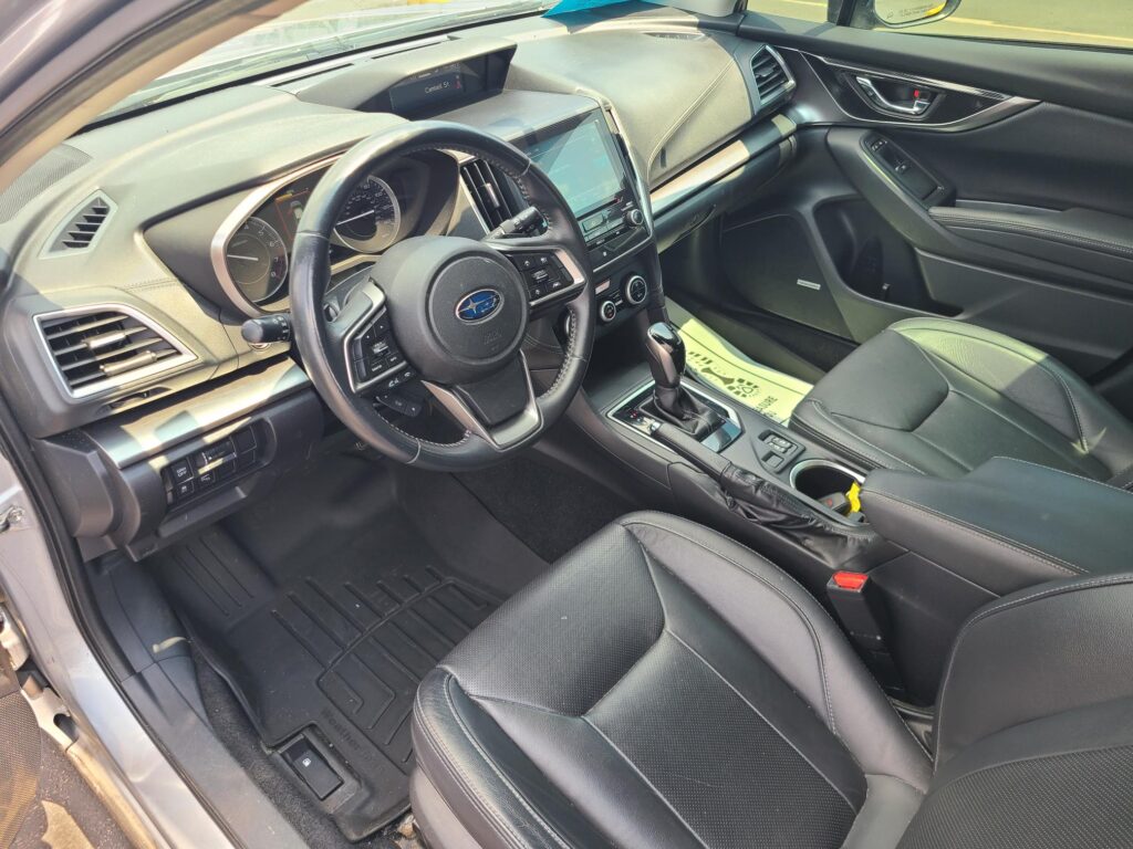 Interior of a modern car cabin showing the driver's seat, steering wheel with a Subaru emblem, and a central touchscreen infotainment system surrounded by control buttons.