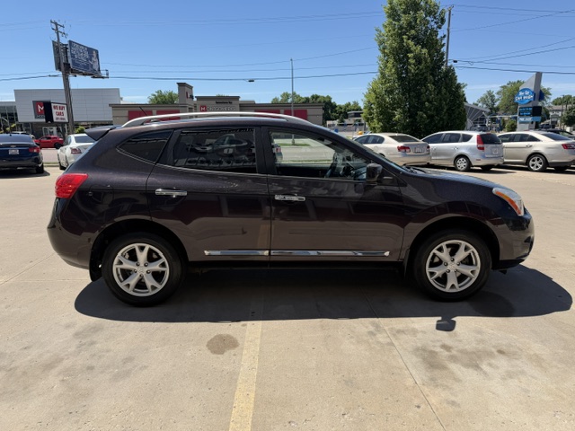 Side view of a dark brown SUV with chrome trim and silver wheels parked in a sunny lot, dealership buildings and other cars in the background.