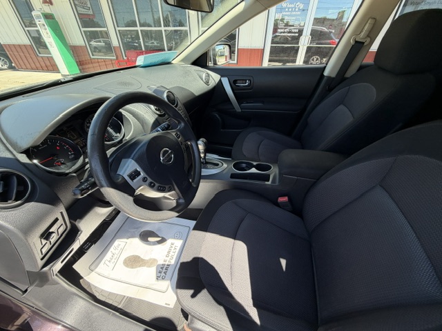 Front interior of a dark-gray car showing the steering wheel, driver and passenger seats, center console with cupholders, and a sunlit dashboard; dealership exterior visible through the windows.