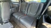 Interior of a SUV showing the black fabric third-row bench with three headrests and visible seat belts, sunlight through the rear window.
